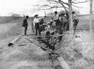 An old photograph of group of men dropping a large pipe into a shallow trench using wooden planks as levers. Early pipelines were buried underground in an attempt to protect them from sabotage by Teamsters.